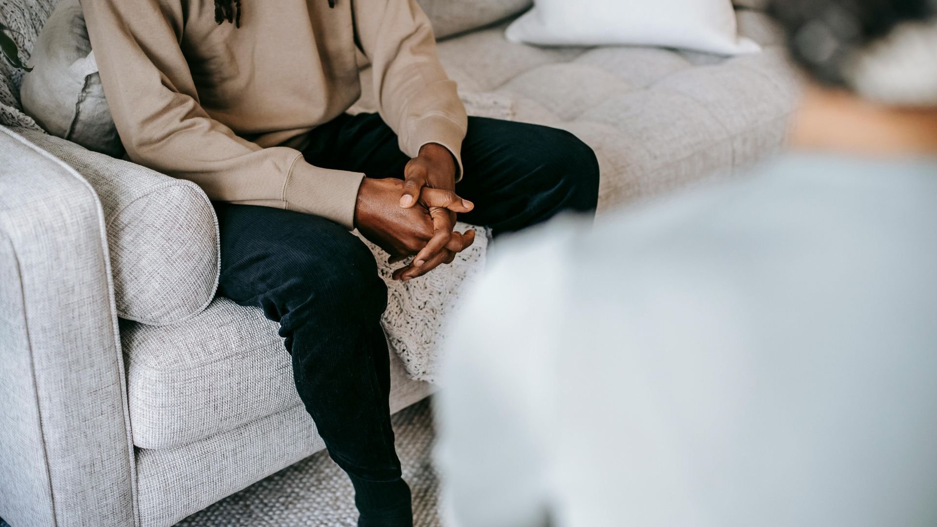 Person sitting on couch, hands clasped, wearing beige sweater and black pants