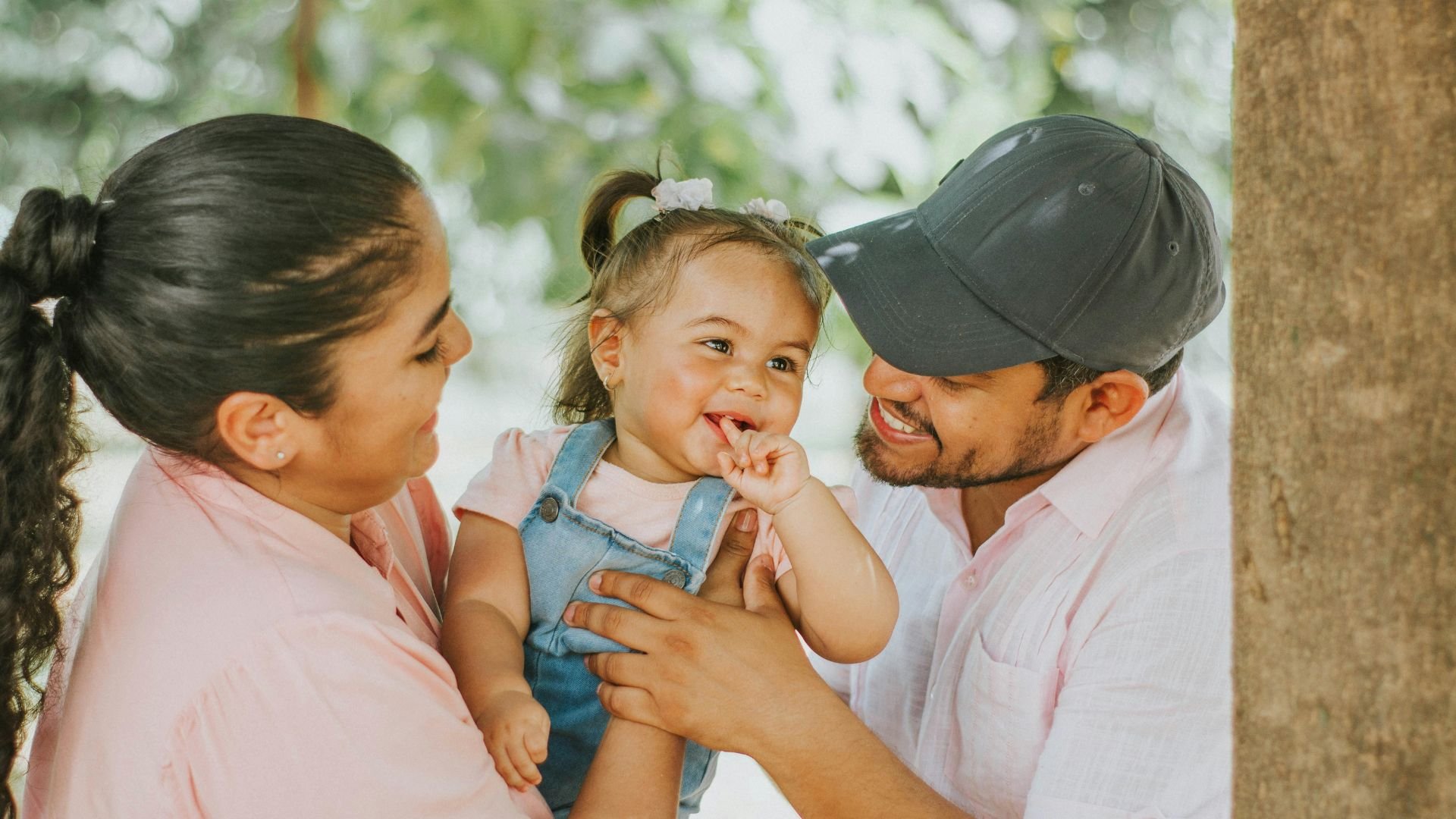 Happy toddler in denim overalls smiling between parents outdoors