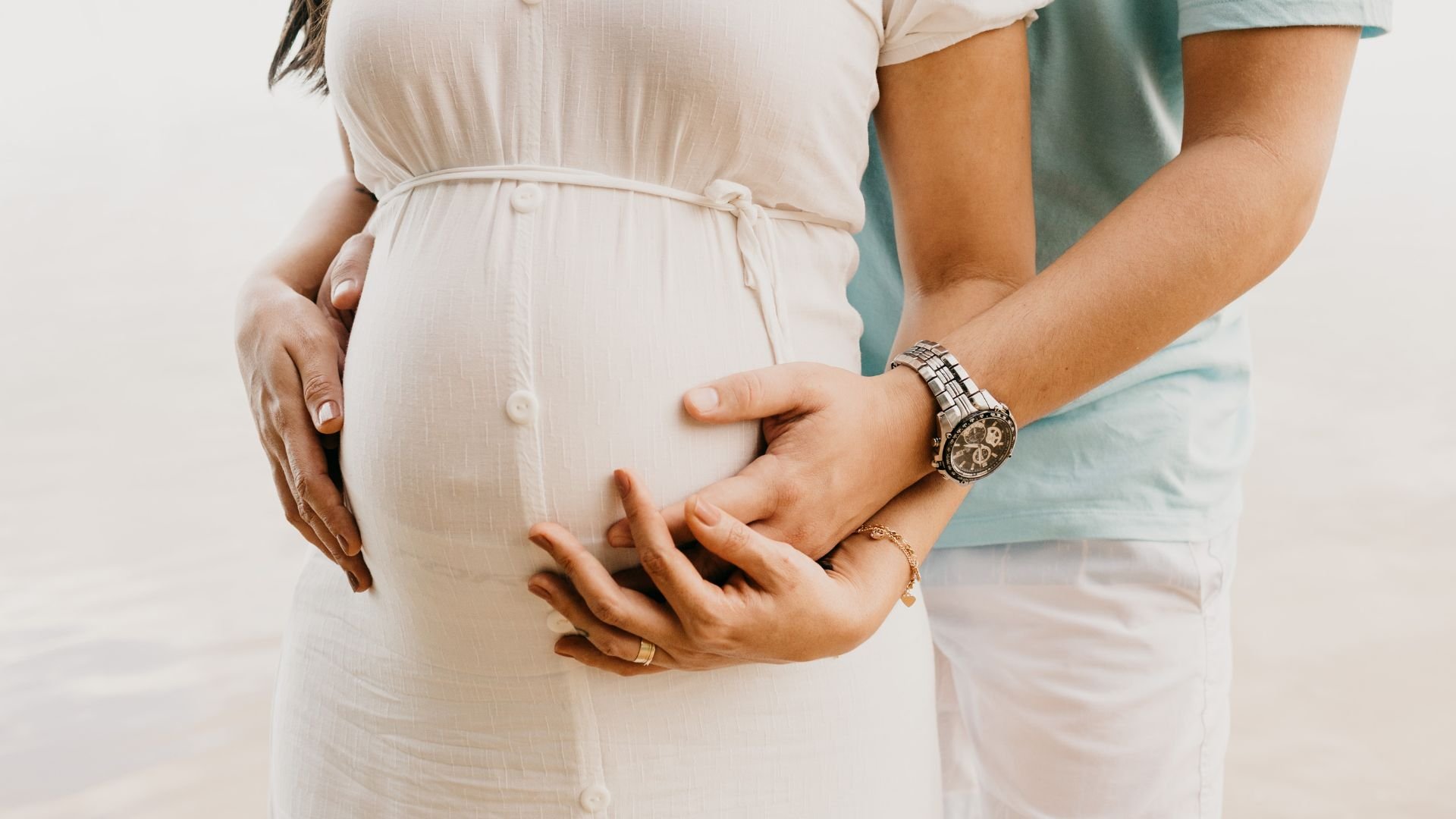 Pregnant woman in white dress with partner's hands on her belly