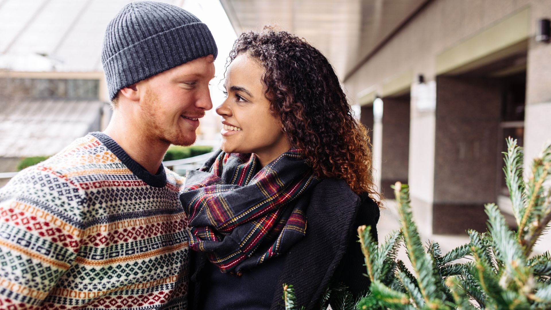 Smiling couple in winter clothing embracing near evergreen plants
